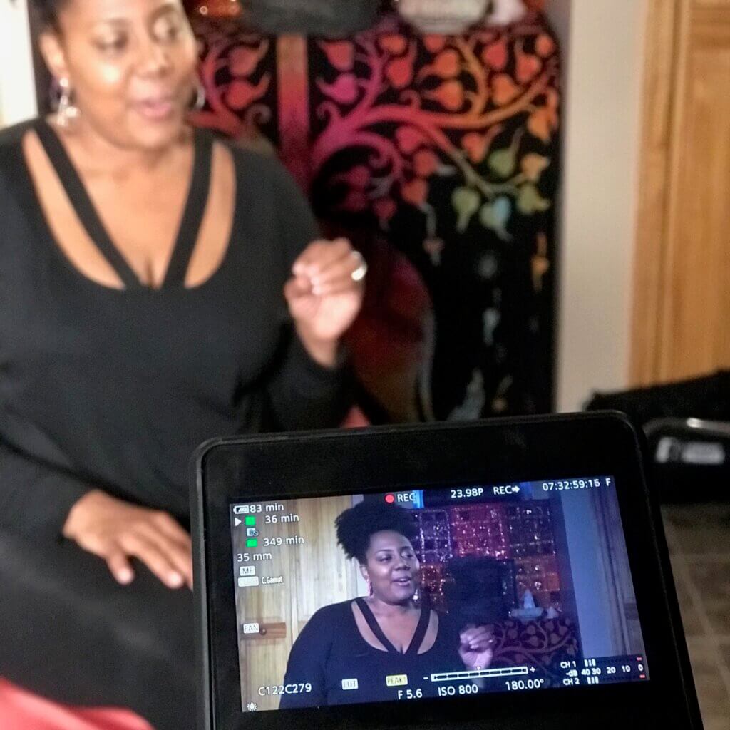 Cherylle, a black woman with her dark hair tied up in a ponytail, speaks to the video camera that is recording. She’s wearing a black top and black pants, and hoop earrings. The background is a fabric colorful tree silhouette.