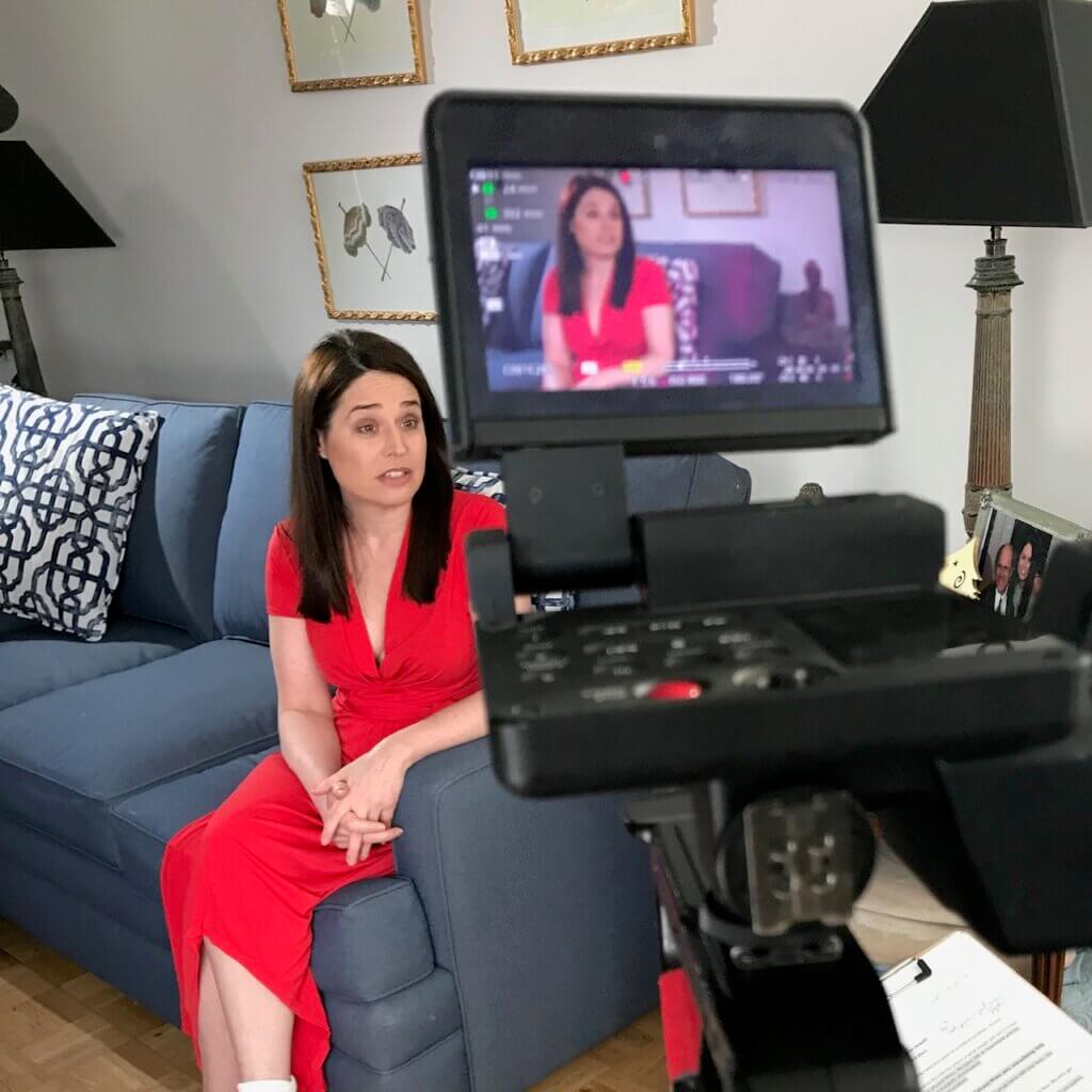 Terri Wilder, a light skinned woman with medium length brown hair and brown eyes, speaks to the video camera that is recording. She’s wearing a bright red dress and sitting on a blue couch, her fingers interlaced on her lap.