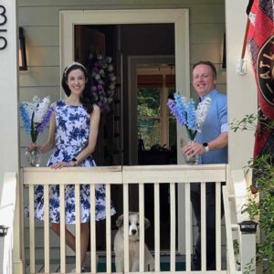 Light-skinned man and woman pose with flowers on a porch. A dog stands between them.