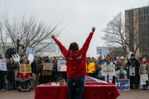 A woman in a red hoodie pumps her fists in the air before a crowd of fellow rally-goers holding their signs of protest in front of them.