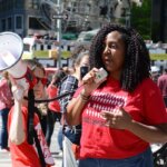 A woman in a red shirt passionately speaks into a megaphone, conveying a message to a gathered crowd.