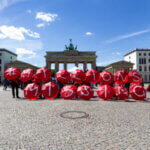 Group holding red umbrellas spelling "MILLIONS MISSING" in front of Brandenburg Gate on a sunny day.