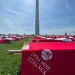 Close up of red blanket draped over a cot. The blanket says "STILL HERE STILL SICK." Hundreds of identical cots dapple the lawn in rows ahead of the Washington Monument under a clear sky.